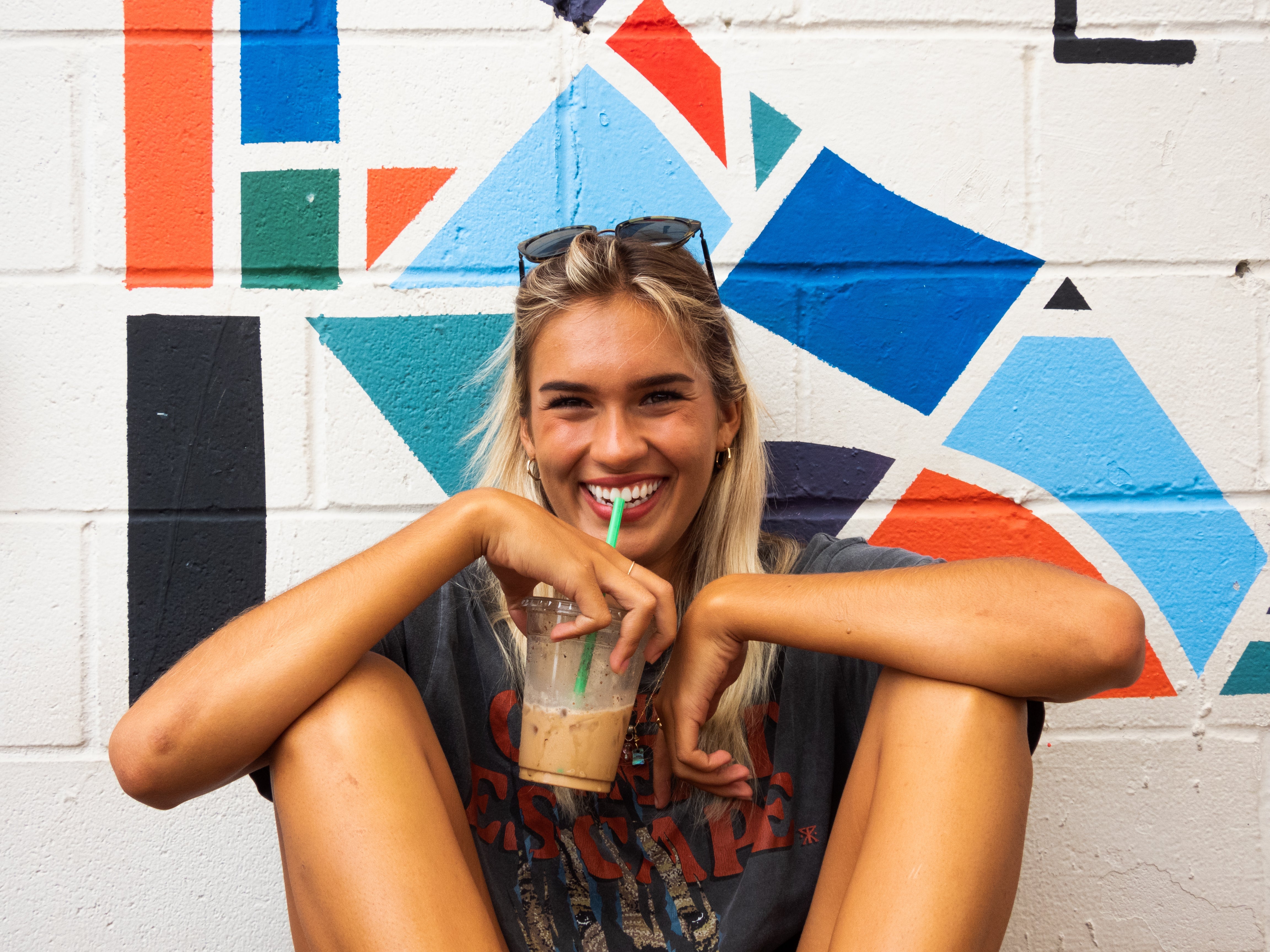 Girl drinking a drink and laughing sitting in front of a colorful wall